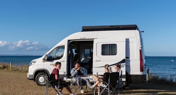 Een vriendengroep zit voor de Mighty Lite DeLITE camper met uitzicht op de zee in Nieuw-Zeeland