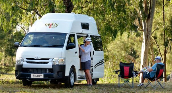 Een vrouw staat bij de open zijdeur van de Mighty Lite DeLITE camper in Australië terwijl haar man in een campingstoel zit