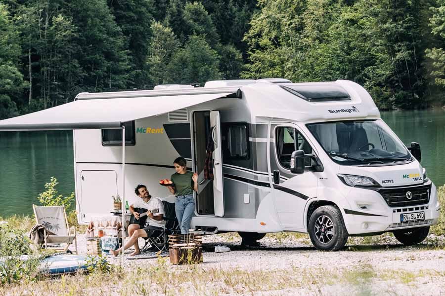 Een vrouw stapt uit de McRent Family Standard camper met een bord eten waar haar man gitaar zit te spelen onder de luifel