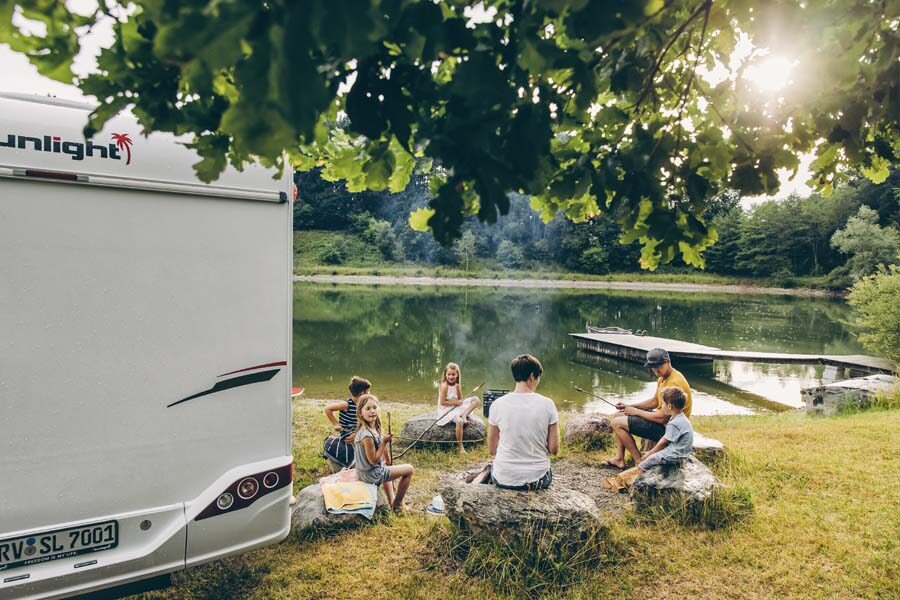 Een gezin zit op stenen naast de McRent Family Plus camper met uitzicht op een meer