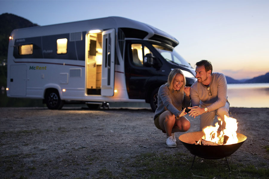 Een koppel staat bij een kampvuur tijdens schemering aan de voorkant van de McRent Comfort Plus camper