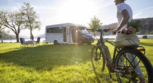Een man loopt met zijn fiets richting zijn vrouw bij de McRent Comfort Plus camper die geparkeerd staat op het gras naast een meer