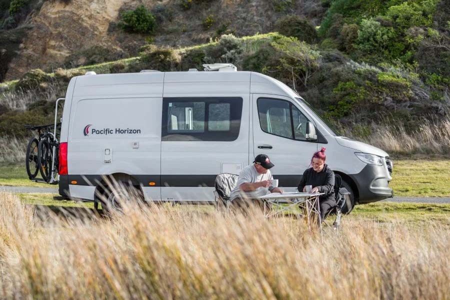 Een stel zit aan tafel op de campingstoelen aan de zijkant van de geparkeerde Pacific Horizon 2 plus 1 camper