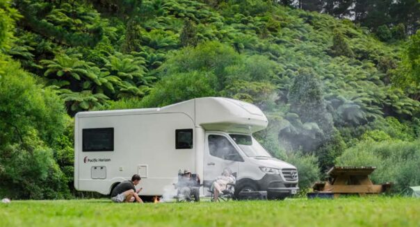Een gezin zit voor de Pacific Horizon Gem 4 camper op de campingstoelen met een zelfgemaakt kampvuurtje in de groene natuur van Nieuw-Zeeland
