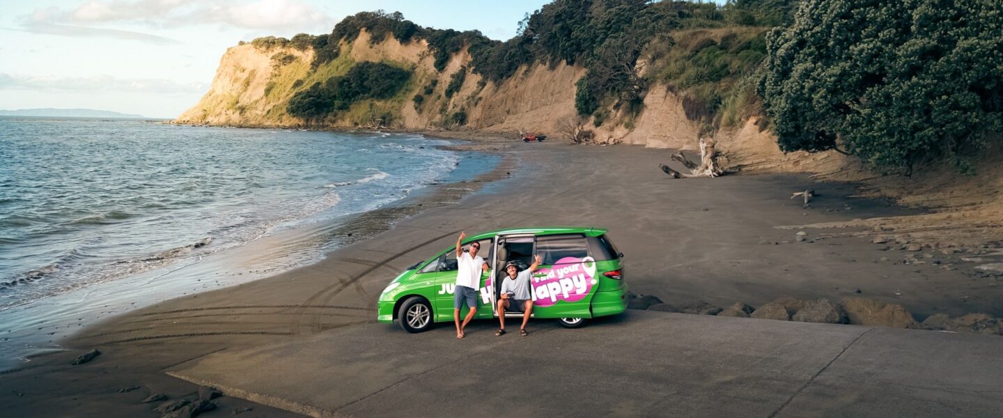 Twee mannen zitten in de deuropening van de Jucy Crib en zwaaien, camper staat geparkeerd op een zwart strand