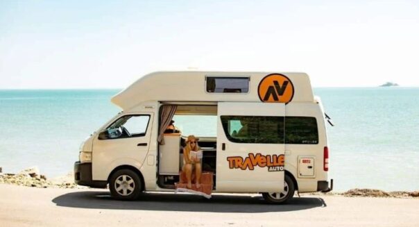 Vrouw leest een boek in de open zijdeur van de Travellers Autobarn Hi5 die geparkeerd staat aan het water