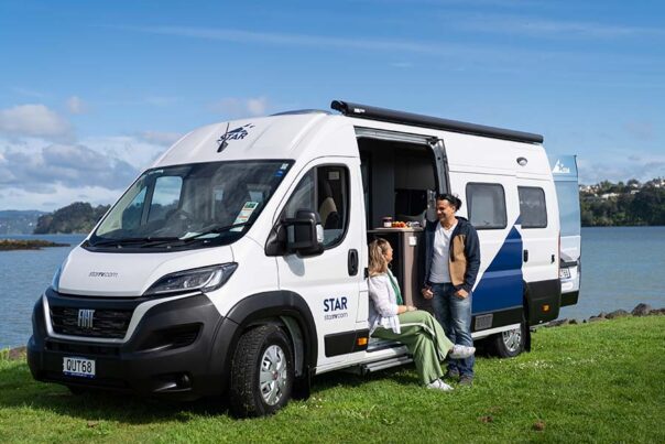 Een vrouw zit in de deuropening van de Star RV Polaris 2 camper die geparkeerd staat naast een meer