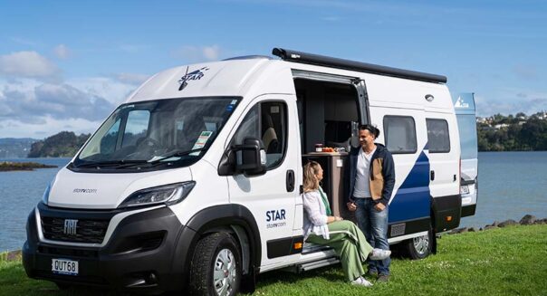 Een vrouw zit in de deuropening van de Star RV Polaris 2 camper die geparkeerd staat naast een meer