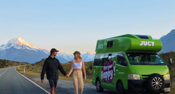Een koppel loopt hand in hand naast een weg en de geparkeerde Jucy Condo camper in Nieuw-Zeeland