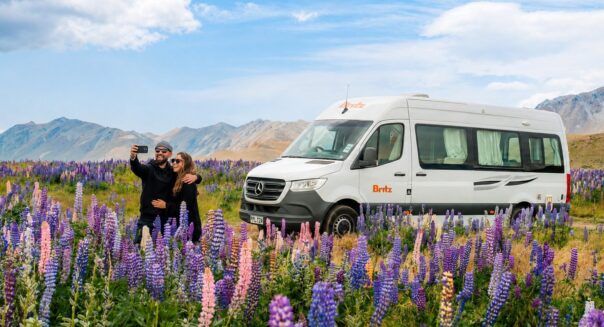 Stel maakt een selfie voor een Britz camper met lupines op de voorgrond