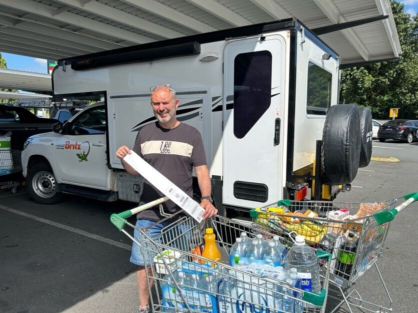 Peter laat de kassabon zien van de eerste camperboodschappen bij 2 volle boodschappenkarren en de Britz Maverick op de achtergrond