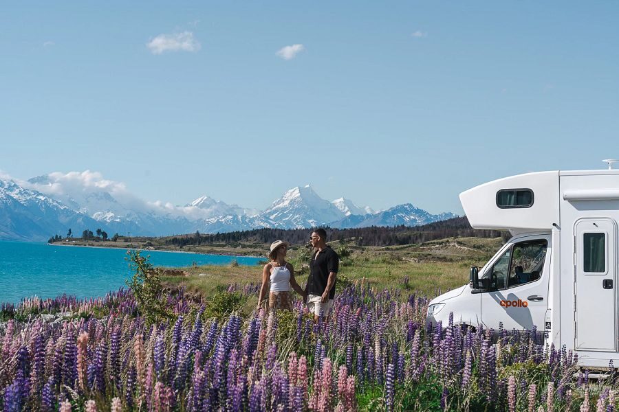 Koppel staat tussen de lupines bij een meer op het Zuidereiland, hun Apollo camper staat geparkeerd