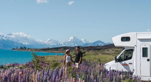 Koppel staat tussen de lupines bij een meer op het Zuidereiland, hun Apollo camper staat geparkeerd