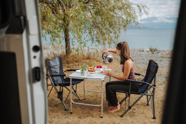 Vrouw schenkt water uit de fluitketel in een mok aan de campingtafel buiten, gezien vanuit de camper
