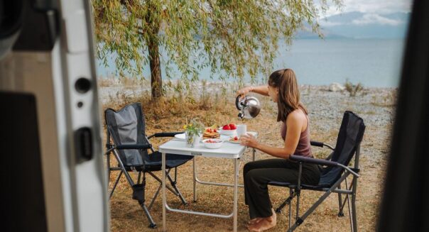Vrouw schenkt water uit de fluitketel in een mok aan de campingtafel buiten, gezien vanuit de camper