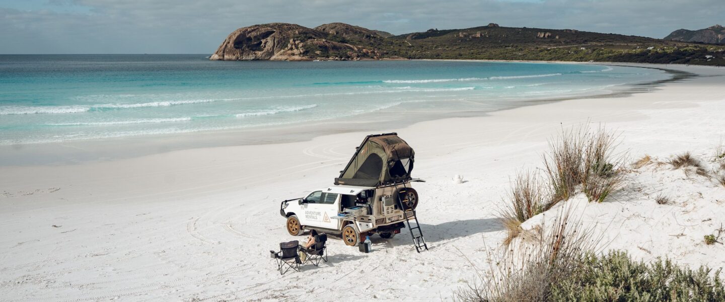 Een 4WD camper van Adventure rentals staat geparkeerd op het witte strand met een vrouw in een campingstoel ernaast