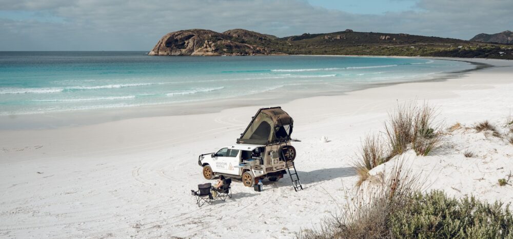 Een 4WD camper van Adventure rentals staat geparkeerd op het witte strand met een vrouw in een campingstoel ernaast
